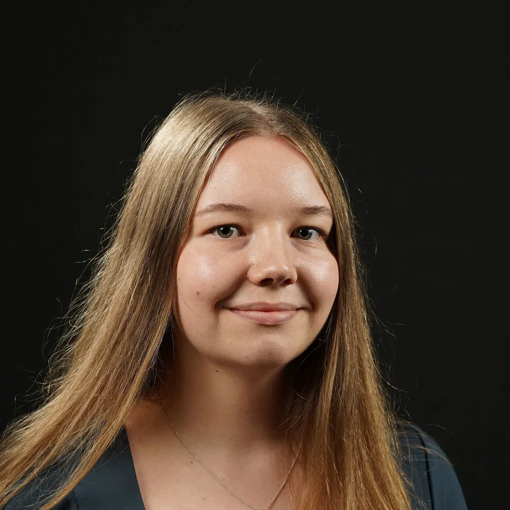 Smiling woman with long, blonde and straight hair against a black background. She wears a dark top and a necklace.