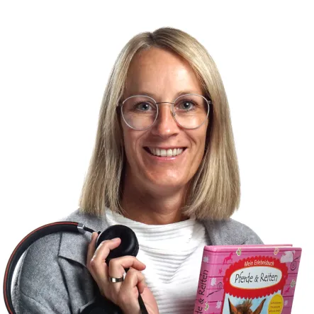 A woman with glasses smiles while holding a pair of headphones and a pink children's book about horses. She wears a gray felt blazer and white shirt.