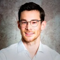 A man with rounded glasses and a smile is wearing a white shirt against a textured gray background. He professionally smiles directly into the camera.