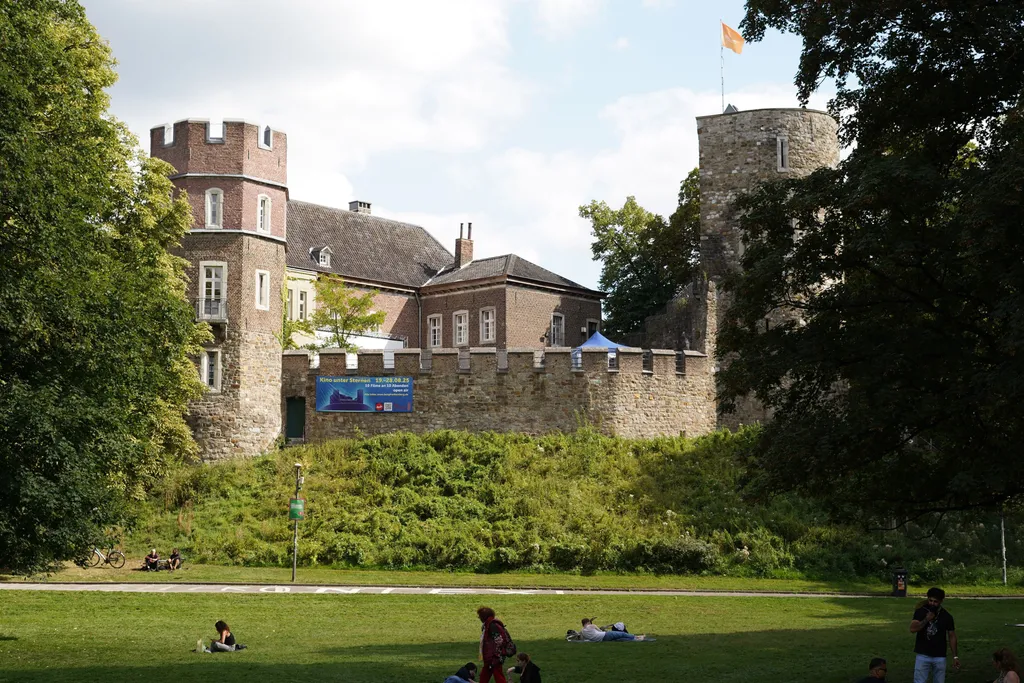 View of Frankenberger Burg in Aachen, a historic stone structure with towers, battlements, and a flag flying atop one turret. People are relaxing on the grassy lawn in front, surrounded by trees and greenery. The scene conveys a peaceful and vibrant atmosphere.