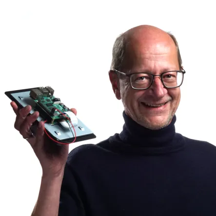 Man holding an electronic device with a visible circuit board, wearing glasses and a black turtleneck, smiling against a white background, conveying a calm and professional tone.