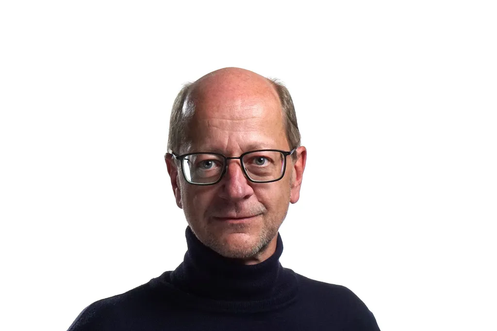 A man with glasses and a partially bald head, wearing a black turtleneck, stands against a plain white background, conveying a calm and professional tone.