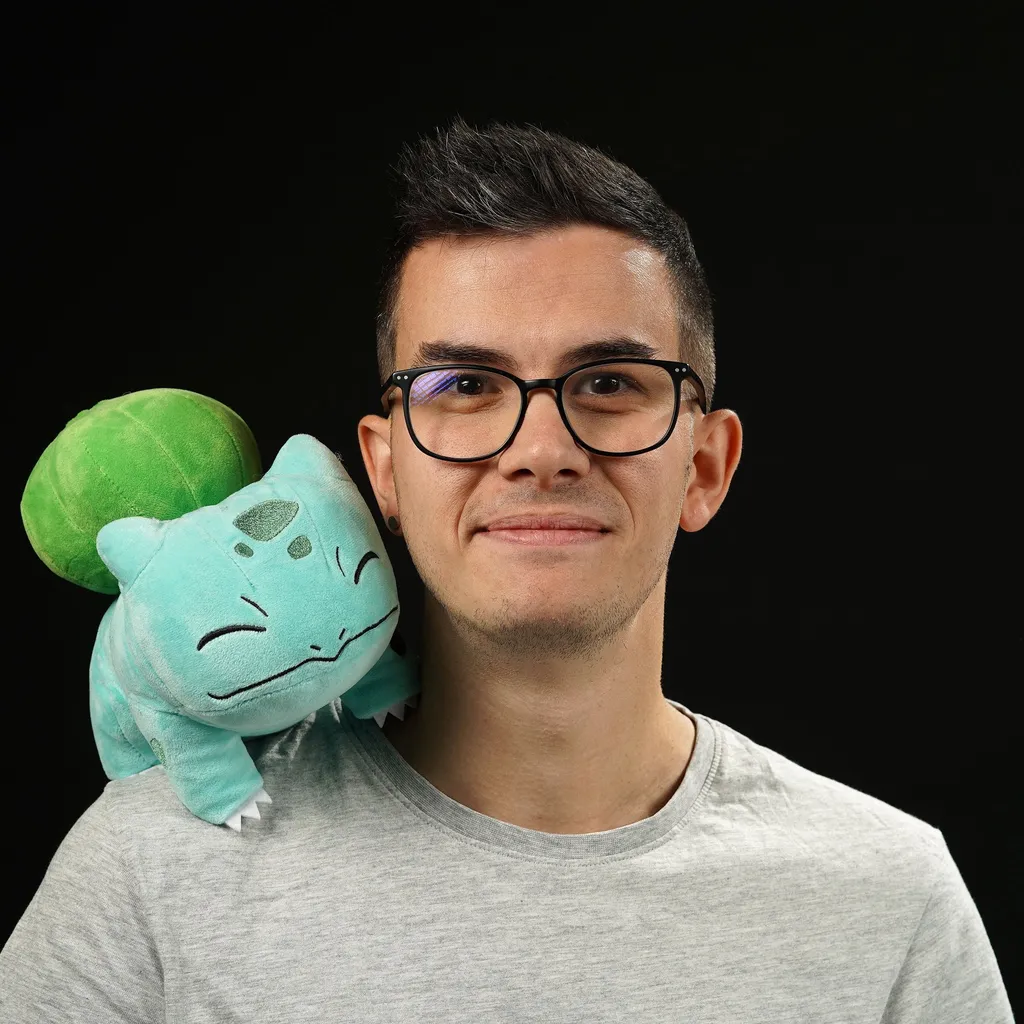 A young man with glasses and a slight smile poses with a plush pokemon toy on their shoulder against a black background, conveying a playful tone.