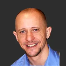A smiling person with a shaved head and light stubble against a dark background, wearing a blue shirt. The tone is friendly and approachable.