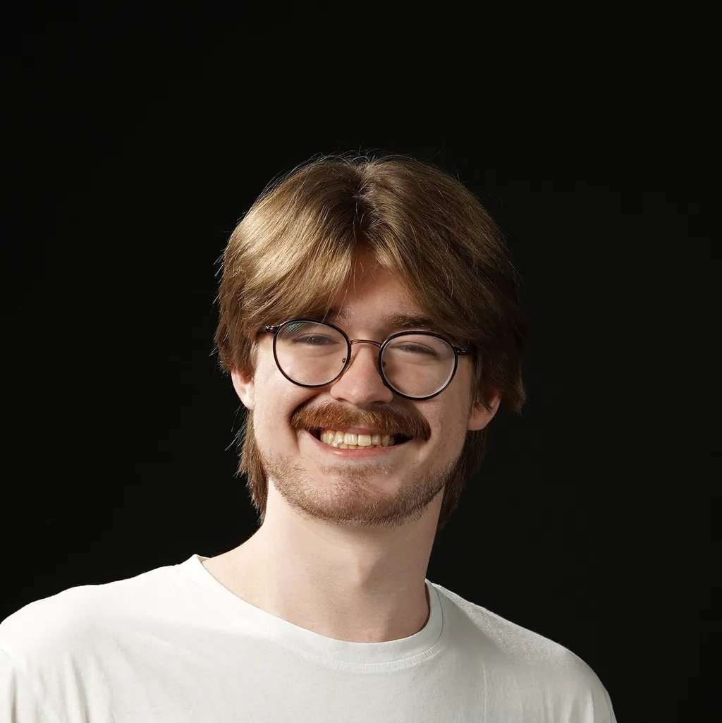Young man with glasses and a mustache smiling warmly against a black background, wearing a white T-shirt, conveying a sense of friendliness.