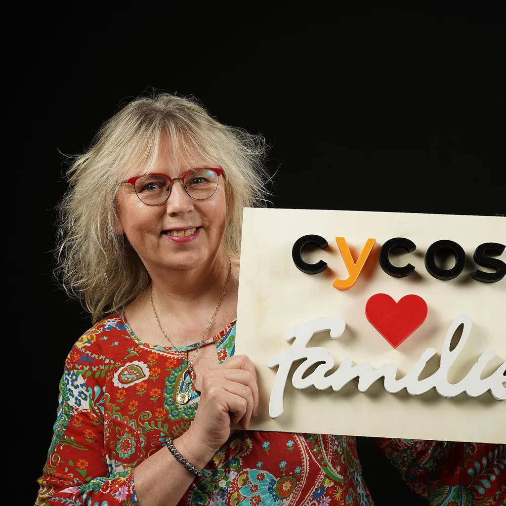 A smiling woman with blonde hair and red glasses holds a sign reading "cycos ♥ Familie" in colorful text. They wear a vibrant, patterned shirt, giving a welcoming look.