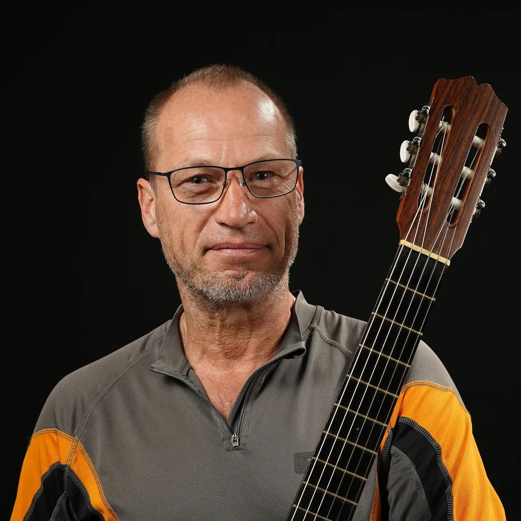 A man with glasses holds an acoustic guitar upright against his shoulder. He wears a gray and orange jacket and stands against a black background.