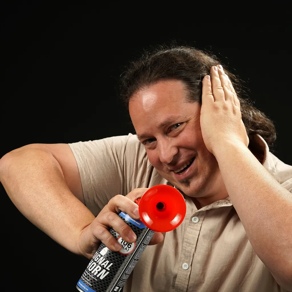 Man with a joyful expression covers one ear while holding a red air horn pointed towards the camera. He wears a beige shirt and has curly brown hair tied back in a ponytail.