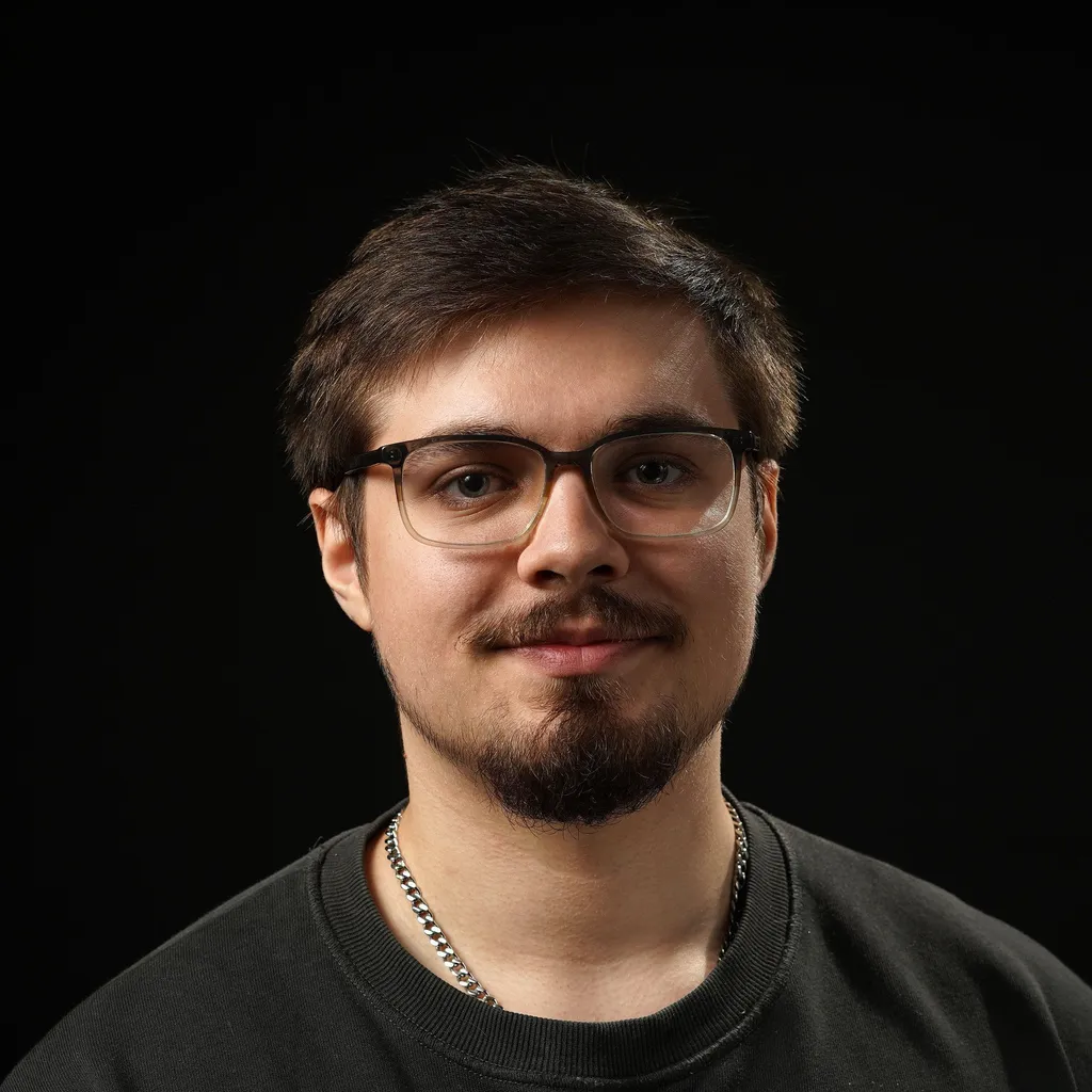 A young man with short brown hair and glasses smiles softly against a dark background. His beard is also brown and cut in a royal shape. He wears a black sweater and a silver chain necklace.