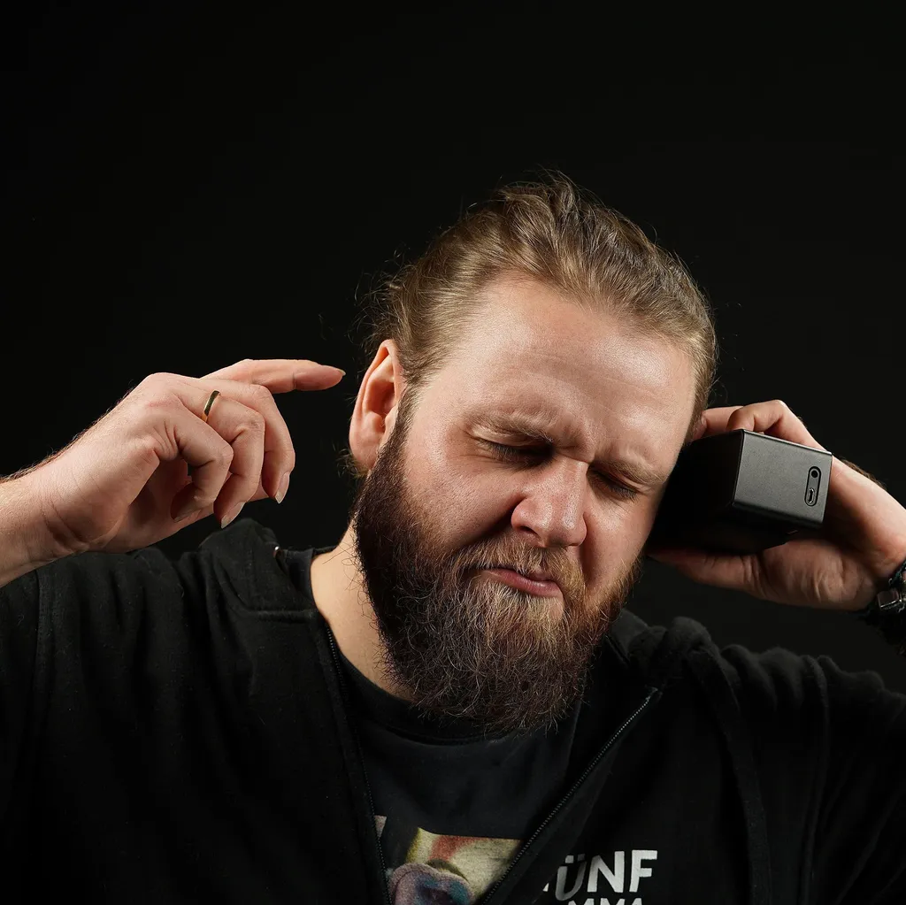 A bearded man in a black shirt holds a speaker to his ear with a tense expression, suggesting movement to rock music. He stands against a dark background, contrasting his dark blonde hair.