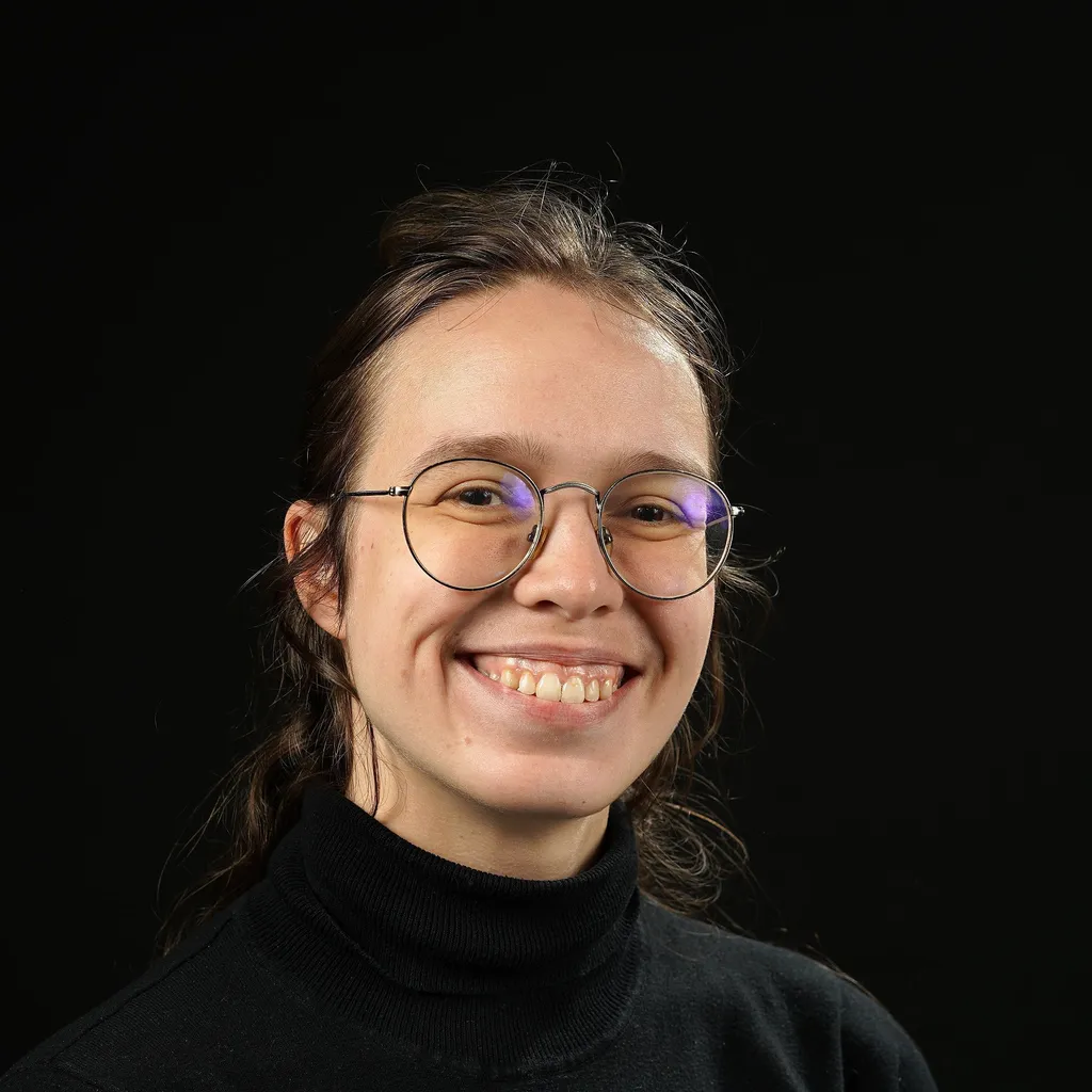 A smiling person with round glasses and dark blonde hair tied back, wearing a black turtleneck against a plain black background. The mood is warm and friendly.