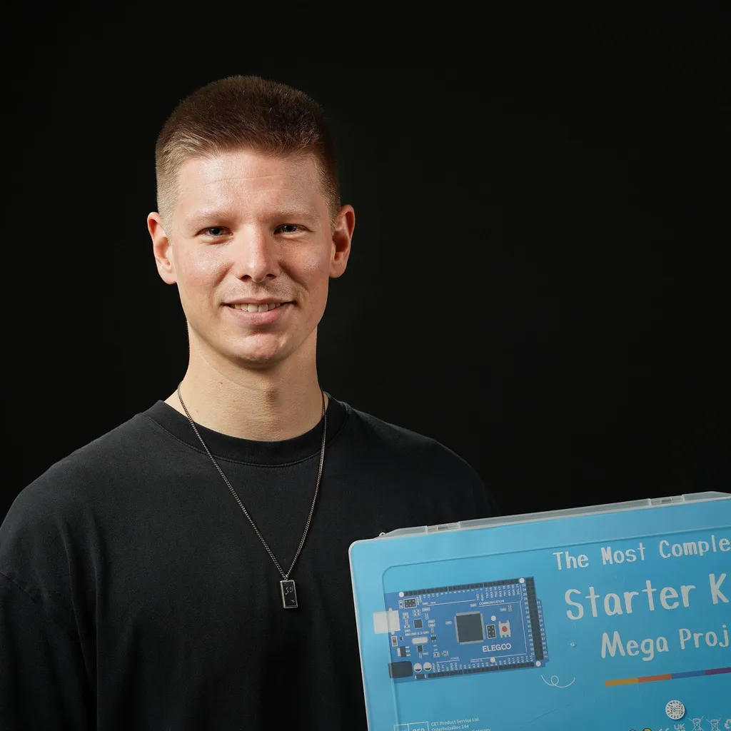 A smiling young man holds an electronics starter kit against a black background. He wears a black shirt and a necklace, conveying enthusiasm and geeky interests.
