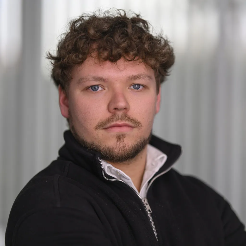 A man with curly hair and a beard looks confidently at the camera. He wears a black zip-up jacket and white shirt. The background is soft-focused gray.