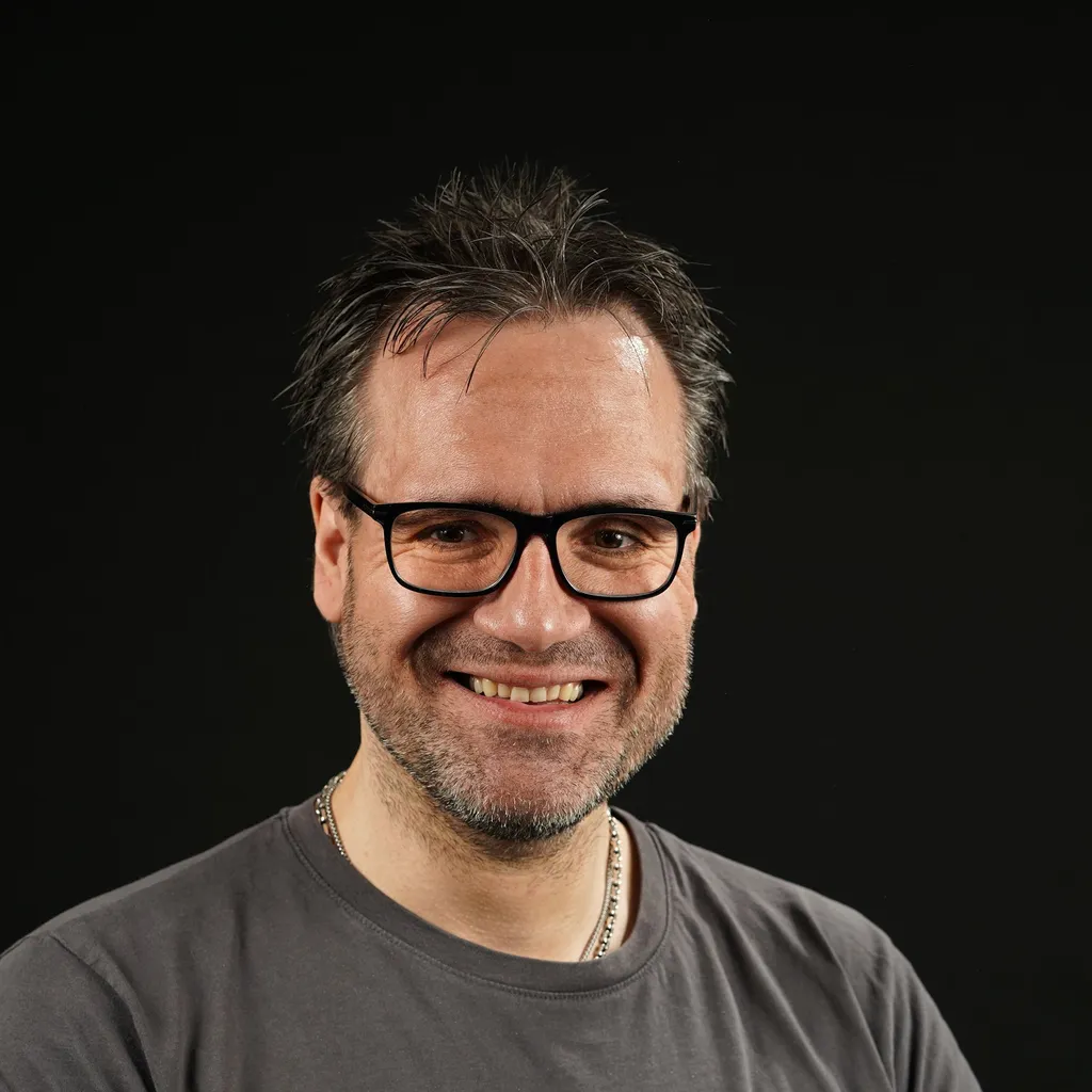 A smiling man with glasses, short hair, and a graying beard, wearing a gray t-shirt. The background is solid black.