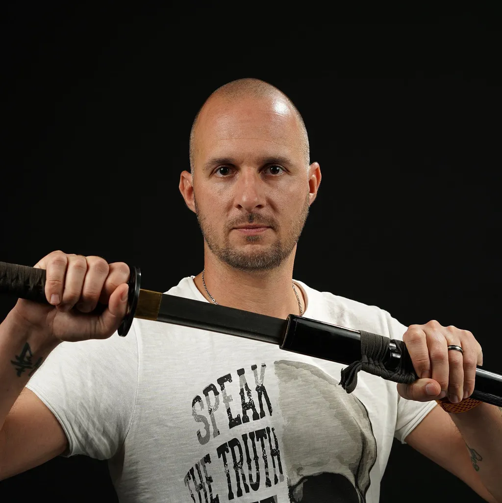 A bald man pulling a katana with one, while holding the scabbard in his other hand. He wears a white shirt with "Speak the Truth" text. He looks determined against a black background.