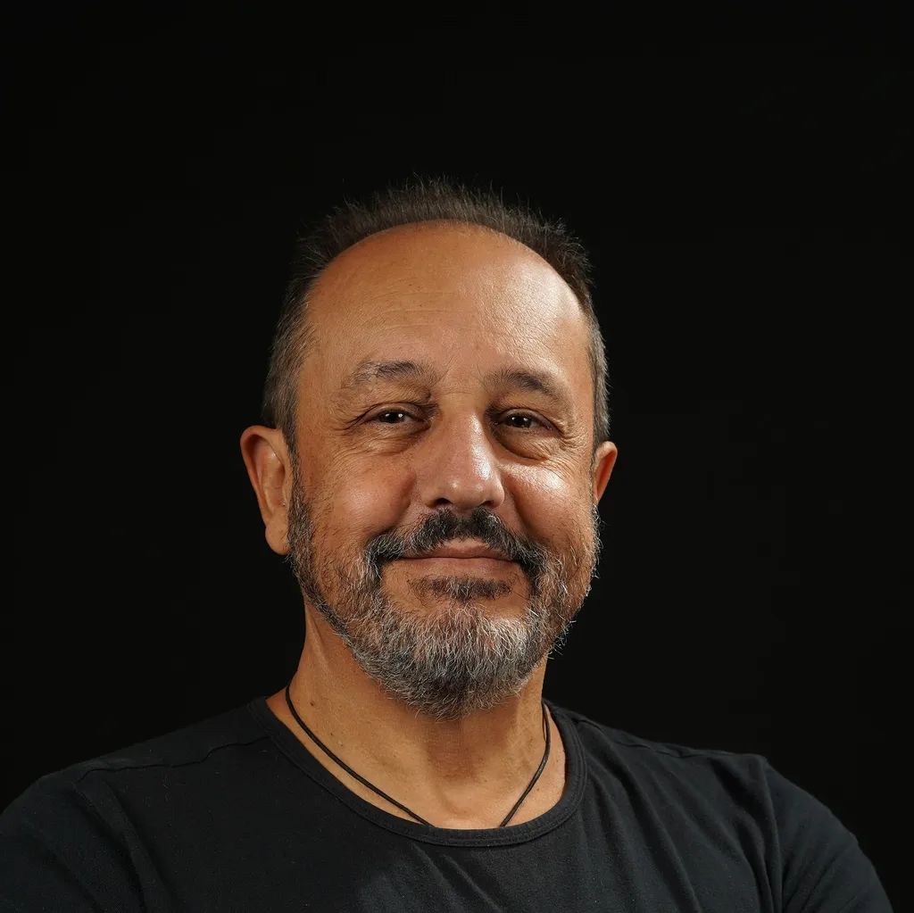 A smiling man with tan skin, a beard and short hair against a black background, wearing a black shirt and a necklace. The mood is warm and friendly.