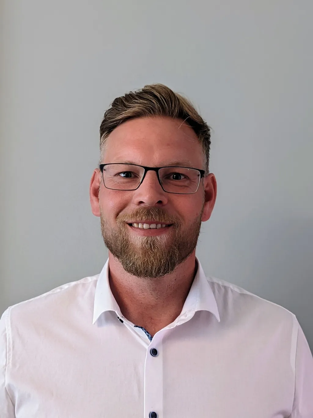 Portrait of a smiling man with glasses, light brown hair, and a beard. He wears a white shirt with dark buttons against a plain gray background.