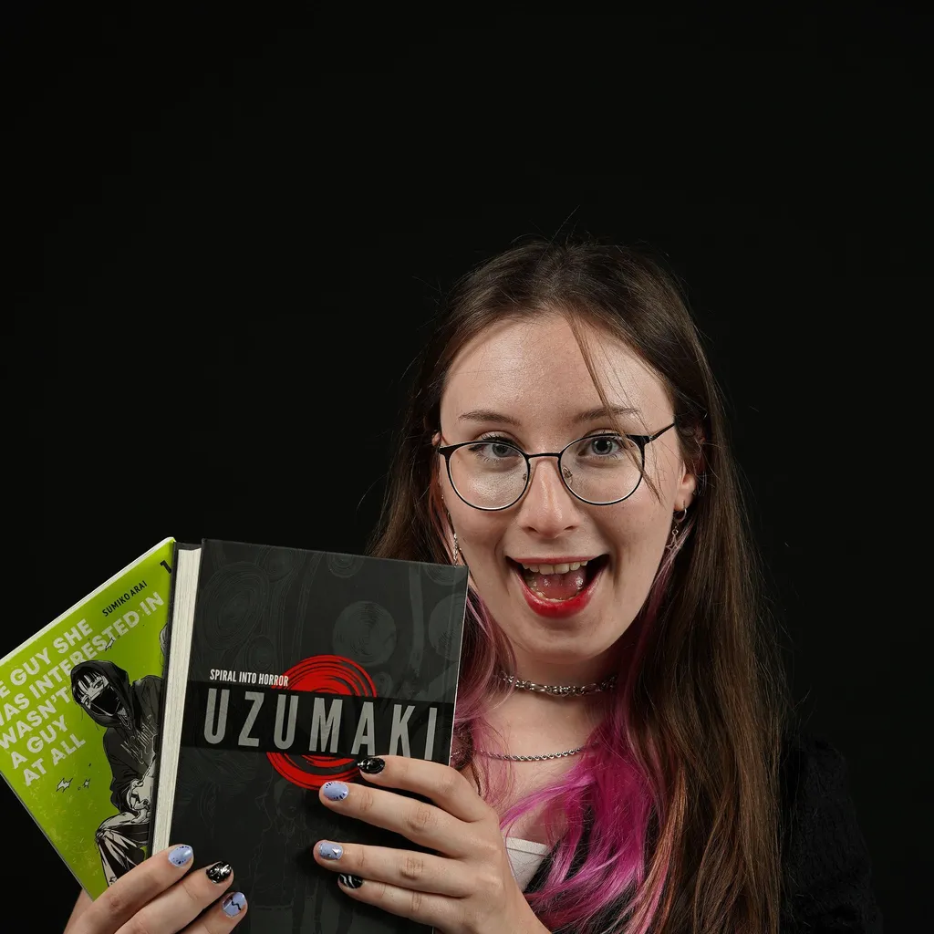 A young woman with glasses and pink streaks in her hair, smiling excitedly and holding two manga books, "Uzumaki" and another with a green cover, against a black backdrop.