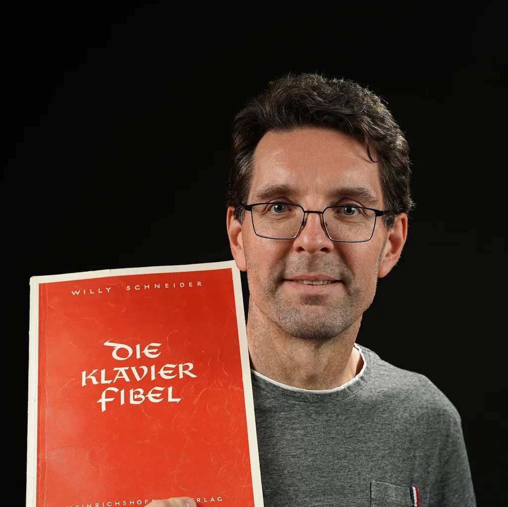 A man wearing glasses and a gray shirt, holding a red book titled "Die Klavier Fibel" against a black background. He smiles slightly.