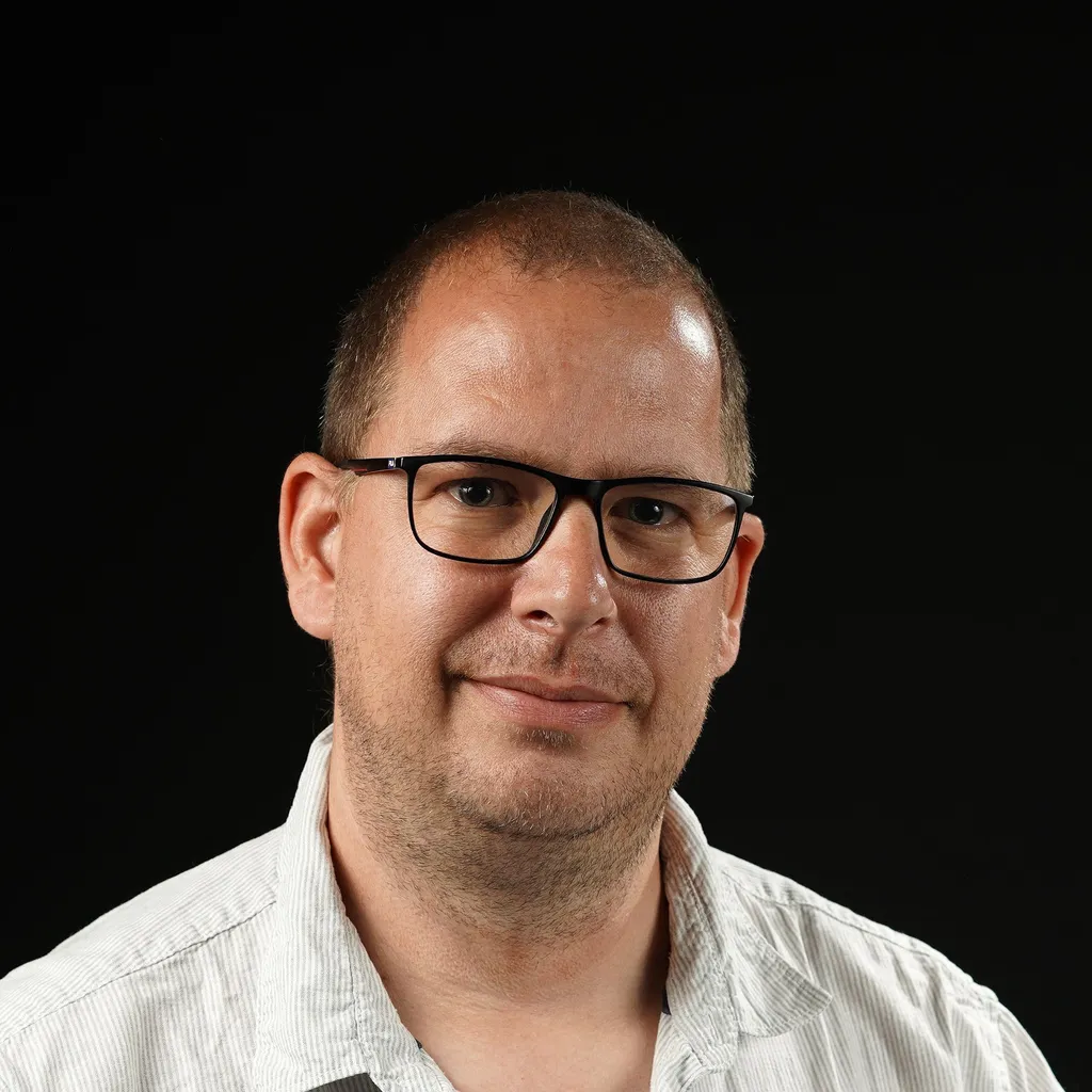A man with a neutral but friendly expression is shown against a black background. He wears a light-colored, pinstriped shirt and has both a shaved head and a stubble. The lighting highlights his face, creating a calm and focused atmosphere.
