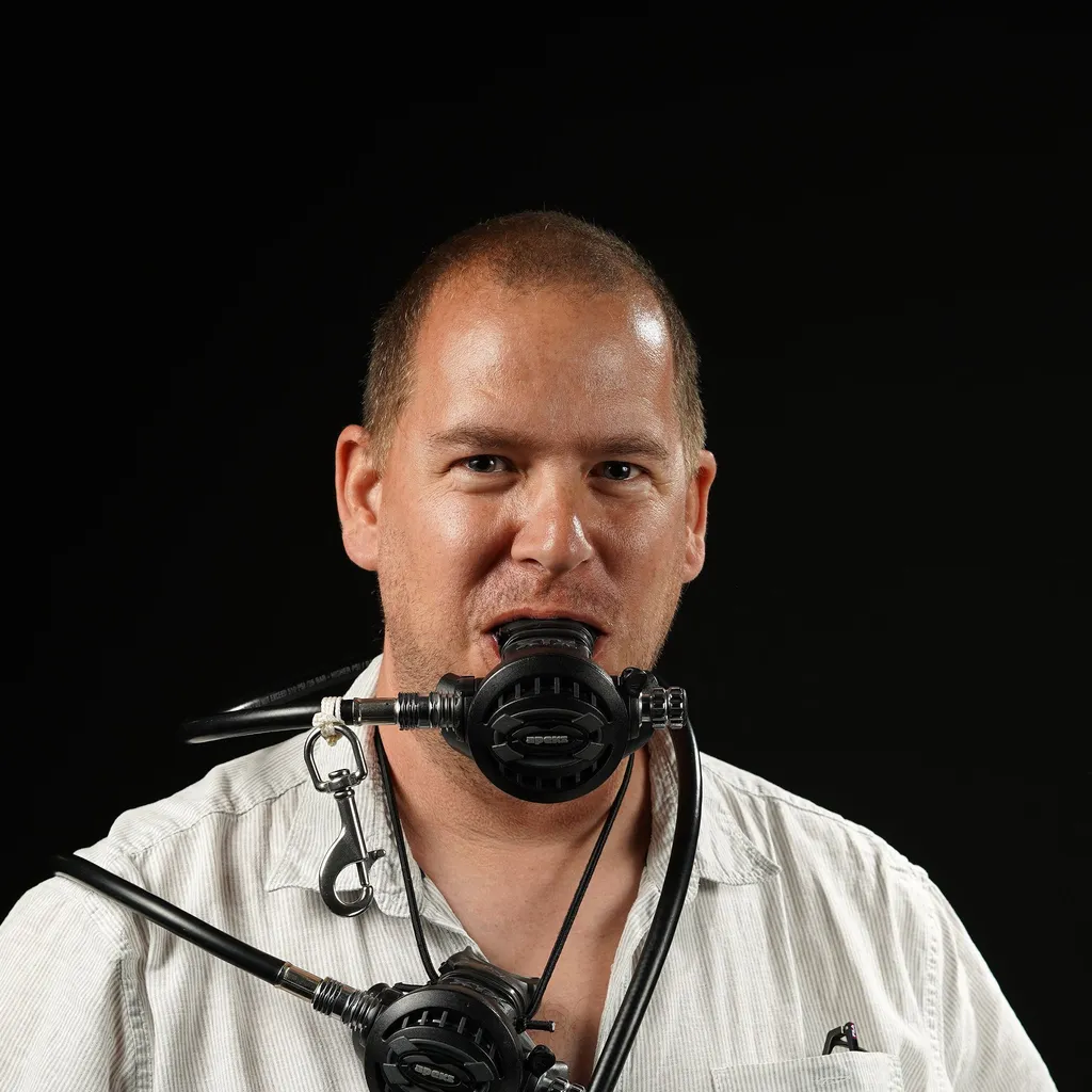 A man against a black background wearing a white shirt holds a scuba regulator in his mouth, with another hanging. While a smile is not visible, he still looks very happy.