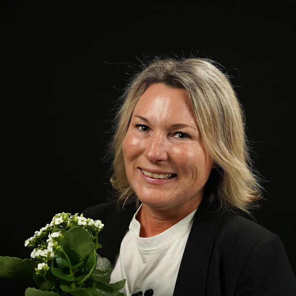 A smiling woman with short blonde hair holds a small plant with white flowers. She is wearing a black jacket over a white shirt, standing against a plain black background.