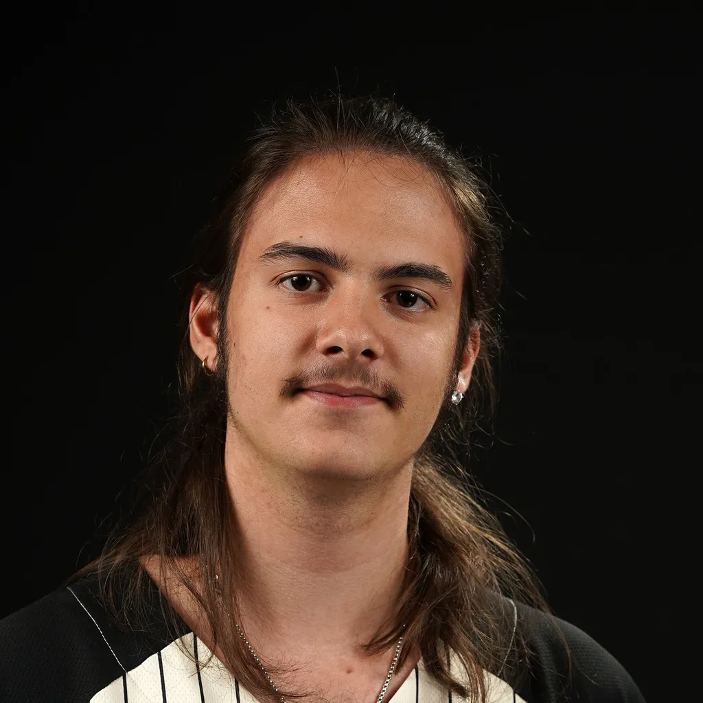 Portrait of a young man with long hair and a mustache, wearing earrings and a striped shirt, set against a dark background. The expression is calm and friendly.