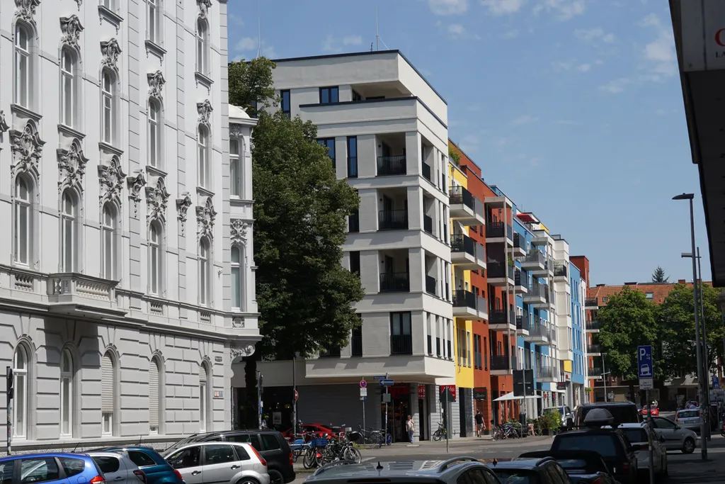 Street view of Viktoriaallee in Aachen, featuring the corner building where cycos is located. The architecture is modern with white facades, black railings, and large windows. Surrounding buildings are painted in vibrant blue, yellow, and red, adding visual contrast. People are walking and biking along the sidewalk, creating a lively neighborhood atmosphere.