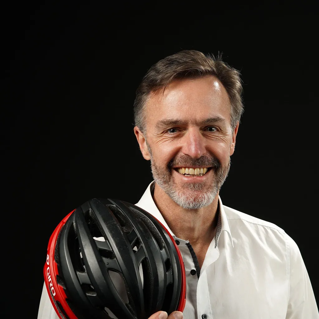 A smiling man in a white shirt with dark buttons holding a black and red bicycle helmet against a dark background. His hair is short and he has a short beard, which is graying. The mood is cheerful and professional.