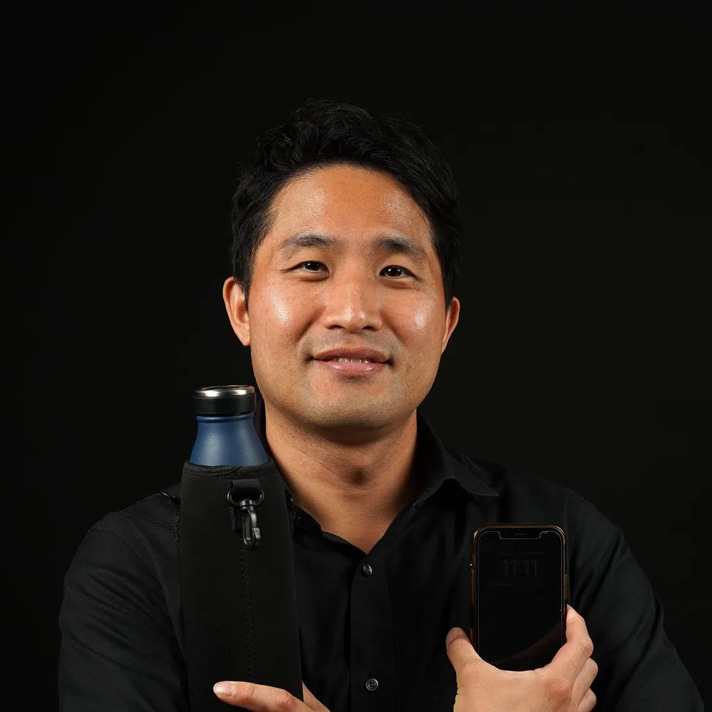 A man holding up a blue water bottle and his phone, while crossing his arms. His skin is warm toned and he has short, straight, black hair. His shirt is black as well and he smiles at the camera.